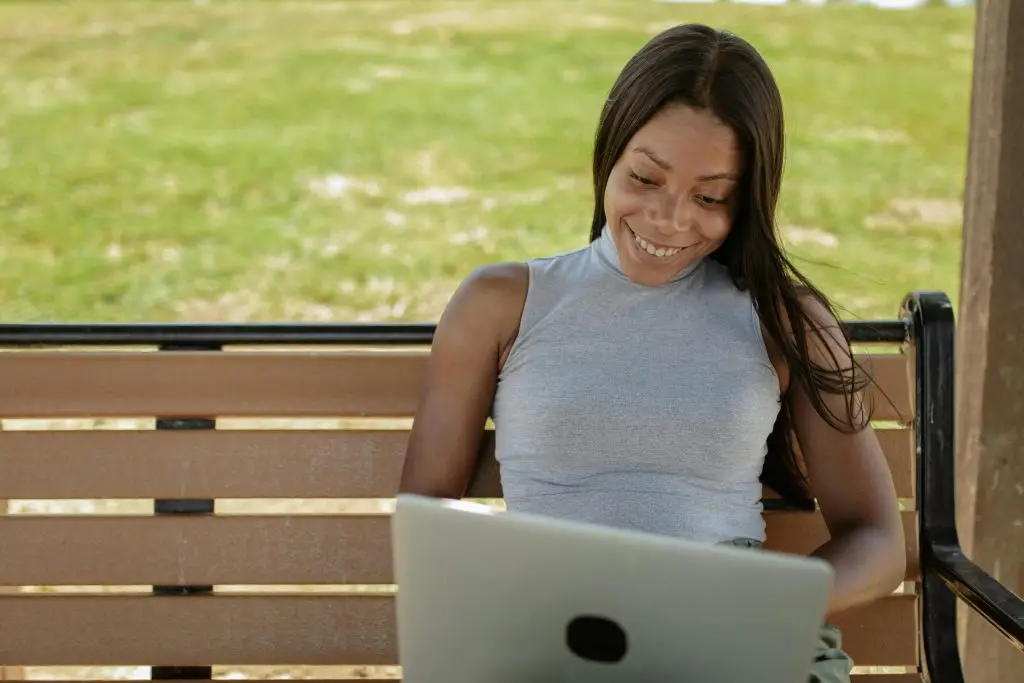 Young woman on a park bench using a laptop and smiling, enjoying remote work outdoors.